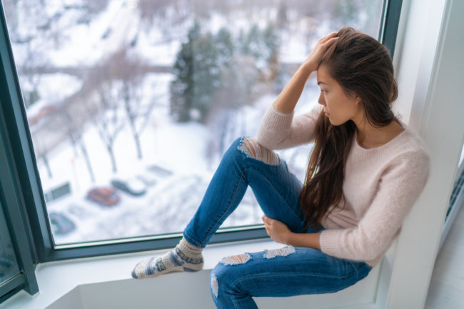 A sad-looking brunette woman with her head in her hand looking out a window at a snowy scene