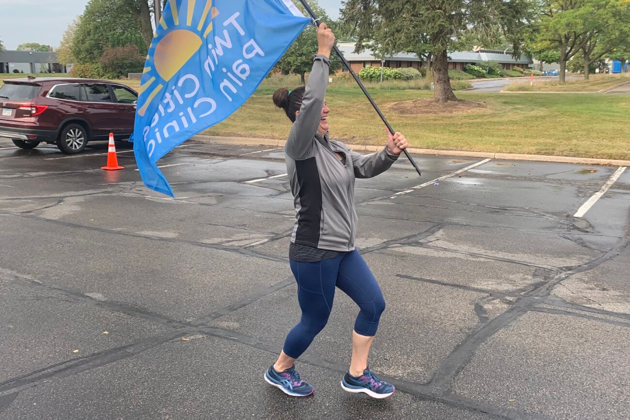 Participant of the Participants running and walking during the 2022 Twin Cities Pain Clinic Pain Awareness 5k running across the finish line with the Twin Cities Pain Clinic flag