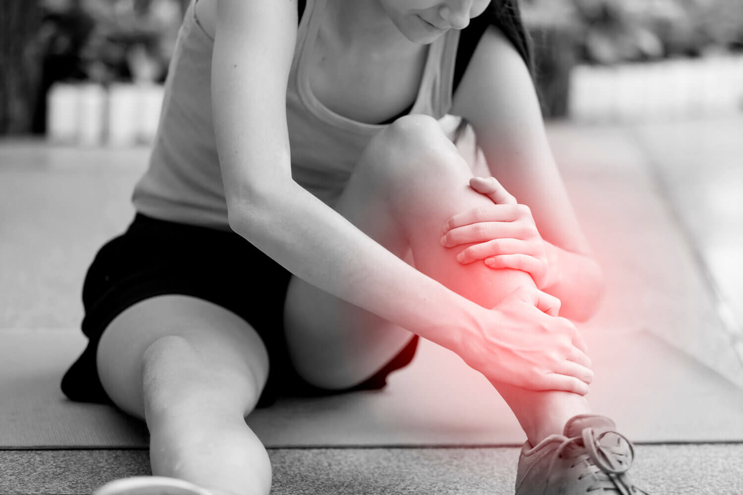 Black and white image of middle age brunette woman sitting on the ground and holding her injured leg which is glowing red to represent extremity pain